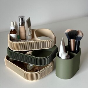 May include: A stack of three ribbed cosmetic trays in beige and olive green, with a separate olive green brush holder. The trays hold various skincare products and the brush holder contains makeup brushes. The items are arranged on a white surface.