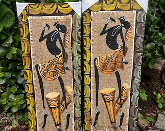 May include: Two framed paintings on canvas with a burlap background. Each painting features a stylized black figure with orange and yellow clothing playing a drum. The paintings are set against a green leafy background.
