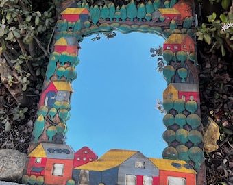 May include: A wooden mirror with a whimsical design featuring colourful houses and trees painted on the frame. The mirror reflects a blue sky.