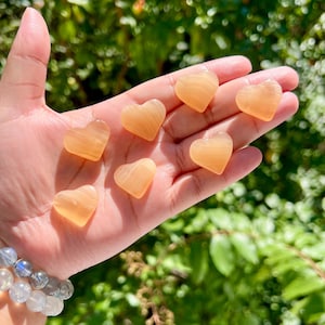 May include: Seven heart-shaped, translucent, orange-yellow stones are held in an open hand. The stones have a smooth, polished appearance and are arranged in a scattered pattern. A bracelet with light blue and clear beads is visible on the wrist.