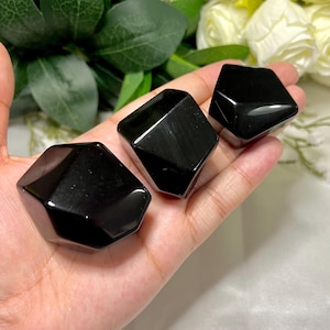 May include: Three polished black obsidian stones with faceted geometric shapes. The stones are held in a hand, with a soft focus background of green leaves and white flowers.