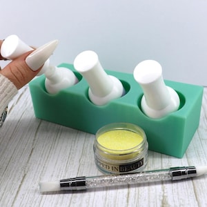 May include: Three white bottles of nail polish in a green silicone holder. A small jar of yellow glitter and a silver and crystal nail art brush are on a wooden surface.