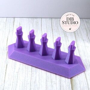 May include: A purple display stand featuring five vertical, faceted posts arranged in a row on a rectangular base. A circular white logo in the upper right corner contains the text "PART OF THE DIB STUDIO COLLECTION" with a small star icon.