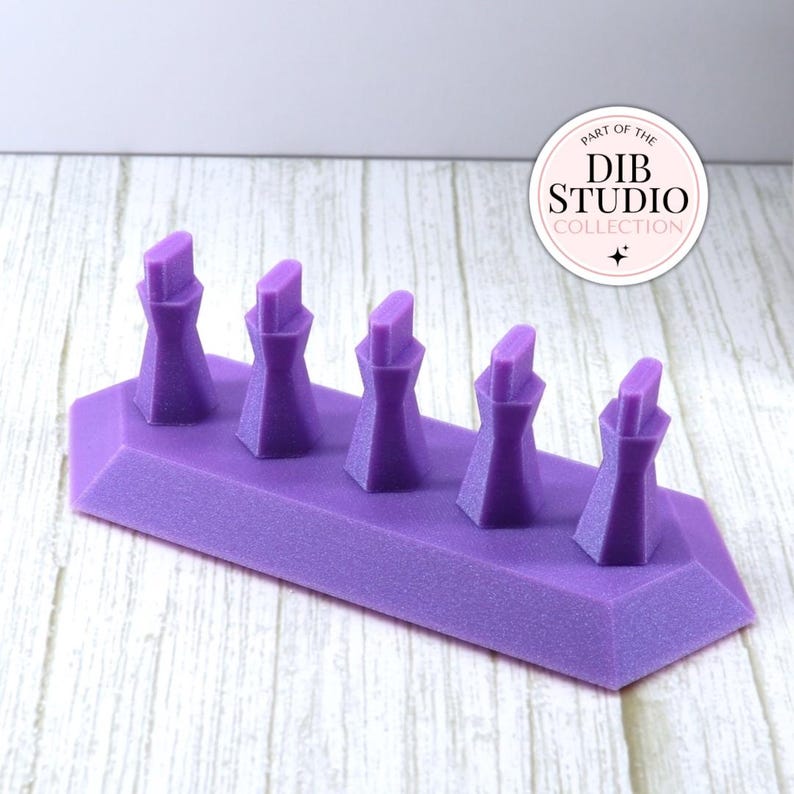 May include: A purple display stand featuring five vertical, faceted posts arranged in a row on a rectangular base. A circular white logo in the upper right corner contains the text "PART OF THE DIB STUDIO COLLECTION" with a small star icon.