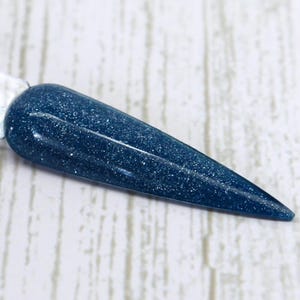 May include: A dark blue nail polish swatch with silver glitter. The polish is applied to a long, pointed nail shape, showcasing the color and sparkle. The background is a light wood grain.