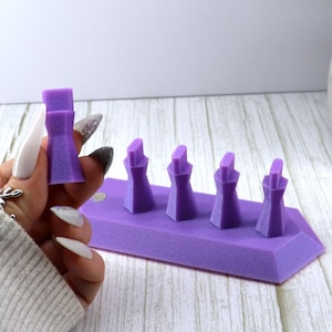 Puede incluir: Un conjunto de cinco soportes púrpuras, geométricos y cónicos con un acabado texturizado. Cuatro de los soportes están unidos a una base rectangular púrpura con extremos angulados, mientras que un soporte idéntico se sostiene por separado.