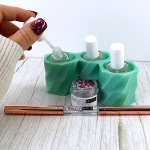 May include: A teal green nail polish holder with three bottles of clear nail polish. A small jar of glitter and a rose gold nail art tool are in front of the holder. The text "Dipped in Beauty" is on the jar and tool.