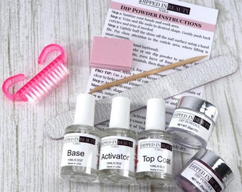 Może przedstawiać: Zestaw do manicure pudrowego z instrukcją, zawierający butelki z bazą, aktywatorem i top coatem, dwa słoiczki z pudrem, pilnik do paznokci, różową szczoteczkę do paznokci i różowy poler. Zestaw zawiera instrukcje.