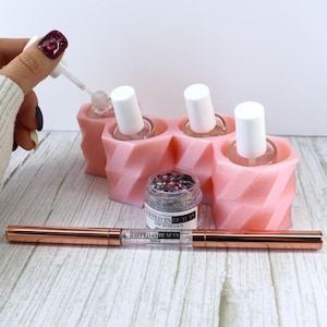 Pode incluir: Um organizador geométrico rosa claro impresso em 3D contendo três frascos transparentes com tampas brancas. Um pequeno frasco de pó de imersão Dipped In Beauty e uma ferramenta de ponta dupla em ouro rosa com o logo Dipped In Beauty Nails.
