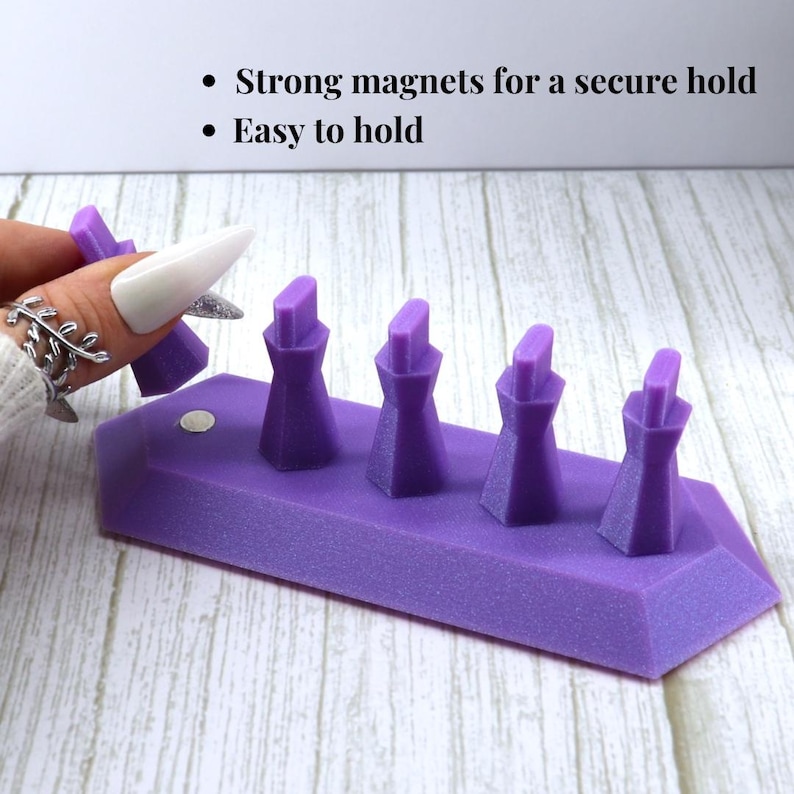 May include: A purple nail art display stand featuring four vertical posts and a magnetic base. The text reads: Strong magnets for a secure hold, Easy to hold. The stand is designed to hold individual nail tips for painting and decoration.