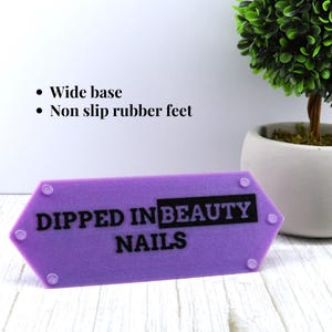 May include: A hexagonal purple acrylic sign featuring the black text DIPPED IN BEAUTY NAILS. The sign includes a wide base and non-slip rubber feet, with four small circular clear fasteners visible on the front surface of the purple acrylic material.