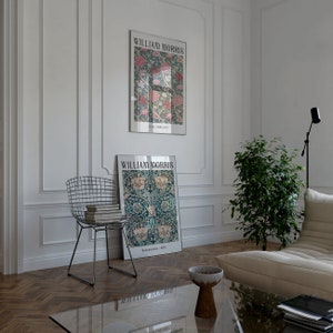 May include: Two framed posters with floral designs and the text "William Morris" on a white wall. The posters are hanging above a wooden floor with a chevron pattern. A metal wire chair with a stack of books sits in front of the posters. A potted plant and a white couch are in the background.