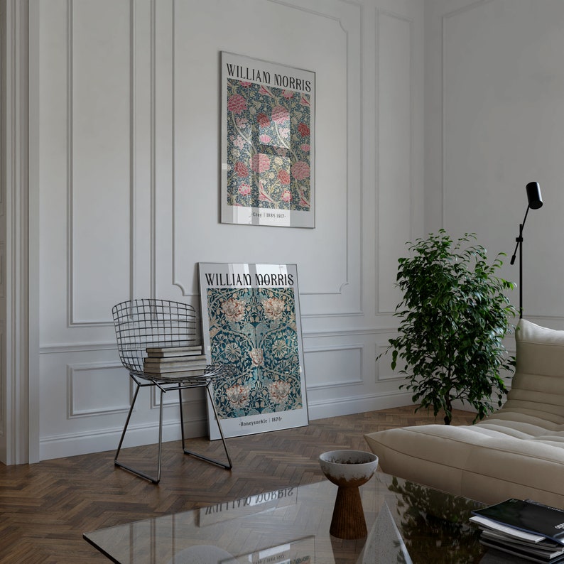 May include: Two framed posters with floral designs and the text "William Morris" on a white wall. The posters are hanging above a wooden floor with a chevron pattern. A metal wire chair with a stack of books sits in front of the posters. A potted plant and a white couch are in the background.