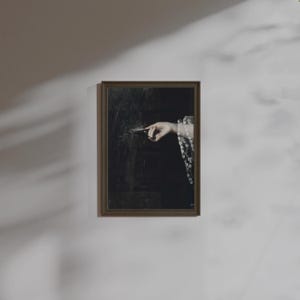 May include: A framed artwork featuring a hand pointing towards a dark, textured surface. The hand emerges from a sleeve with a white floral pattern. The frame is a warm brown color, and the artwork is mounted on a white wall.