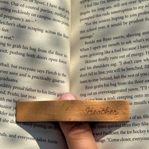 May include: A wooden bookmark with the name "Heather" engraved on it. The bookmark is being used to hold open a book.