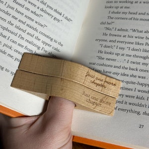 Könnte beinhalten: Ein hölzerner Buchhalter mit dem Text "Just one more chapter..." eingraviert. Der Halter ist so konzipiert, dass er über einen Finger geschoben wird und das Buch offen hält. Das Buch hat orangefarbene Kanten und ist auf einer Textseite geöffnet.
