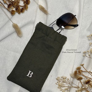 May include: Olive green sunglasses case with a pale mauve thread drawstring closure. The case has a white embroidered letter "B" on the front.