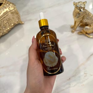 Peut inclure: Une bouteille en verre marron avec un bouchon et un compte-gouttes dorés, étiquetée "Vanilla Icecream Soap" avec une image de crème glacée. La bouteille est tenue dans une main. Un objet décoratif doré est en arrière-plan.