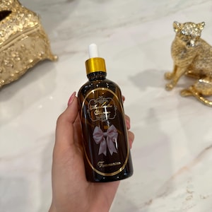 May include: A brown glass bottle with a gold cap and white dropper is held in a hand. The bottle has a label with the text "Marshmallow Blossoms" and a pink bow. The word "Femmenino" is printed at the bottom of the label.