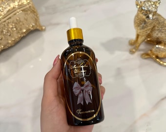 Puede incluir: Una botella de vidrio marrón con tapa dorada y cuentagotas blanco se sostiene en una mano. La botella tiene una etiqueta con el texto "Marshmallow Blossoms" y un lazo rosa. La palabra "Femmenino" está impresa en la parte inferior de la etiqueta.