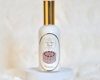 Puede incluir: Un frasco de perfume blanco con tapón dorado y una etiqueta con una ilustración de pastel. La etiqueta dice "Sexy Sugar Vanilla" y "Femmenina" en escritura elegante. La botella se muestra sobre un pedestal blanco.