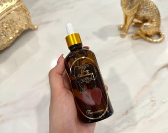 以下が含まれることがあります： 金色のキャップとドロッパーが付いた茶色のガラス瓶で、「Lazy Strawberry」のラベルとイチゴのイラストが描かれています。瓶は手で持たれており、背景には金の猫の像と装飾的な箱があります。