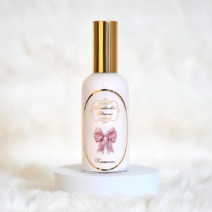 以下が含まれることがあります： 金色のキャップと「Marshmallow Blossoms」と「Femmenina」の文字が入ったラベルが付いた白いボトル。ラベルにはピンクのリボンのグラフィックも含まれています。ボトルは白い台座に展示されています。