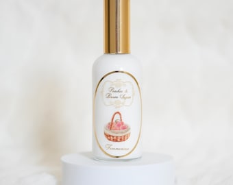 Puede incluir: Un frasco de perfume blanco con tapa dorada y etiqueta decorativa. La etiqueta presenta una cesta de melocotones y el texto "Peaches & Brown Sugar" y "Femmenina" en un marco dorado. El frasco está sobre un pedestal blanco.