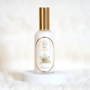 Könnte beinhalten: Eine weiße Parfümflasche mit goldenem Verschluss und einem Etikett mit der Aufschrift "Vanilla Lace". Das Etikett zeigt eine Blumenillustration und das Wort "Temmerina". Die Flasche steht auf einem weißen Sockel.