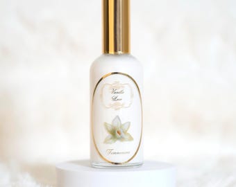 May include: A white perfume bottle with a gold cap and a label that reads "Vanilla Lace". The label features a floral illustration and the word "Temmerina". The bottle is displayed on a white pedestal.