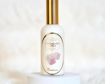 Puede incluir: Una botella blanca con tapa dorada y una etiqueta que dice "Marshmallow Musk" con dos malvaviscos rosas en forma de corazón. La botella está sobre un pedestal blanco sobre un fondo blanco y esponjoso. La palabra "Femmenina" está debajo.