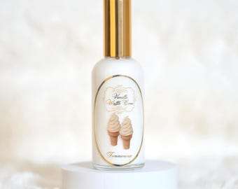 May include: A white perfume bottle with a gold cap, labeled "Vanilla Waffle Cone" with two ice cream cone illustrations. The bottle is on a white pedestal, with a soft, white background.