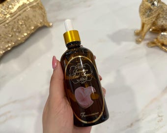 以下が含まれることがあります： 金色のキャップとドロッパーが付いた茶色のガラス瓶が手に持たれています。ボトルには「Seductive Pink Sugar」の文字とピンクの桃のイラストが入ったラベルが付いています。「Femenina」の文字がラベルの下部に印刷されています。