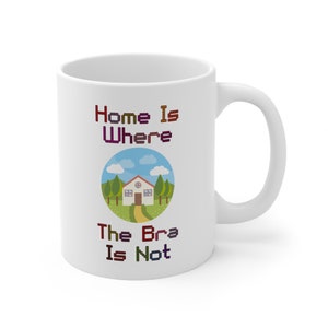 May include: White ceramic mug with a colorful pixelated design that says "Home is where the bra is not" and a cartoon illustration of a house with a green lawn and blue sky.