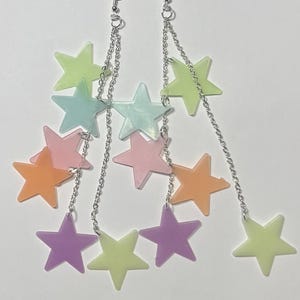 May include: A pair of drop earrings featuring silver-toned metal chains and multiple star-shaped charms in pastel shades of lime green, light blue, soft pink, orange, and purple, suspended from standard silver-toned metal earring hooks.
