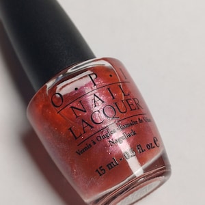 May include: A bottle of OPI NAIL LACQUER in a reddish-orange shade with a black cap. The bottle has text in English, French, and German, and contains 15 ml of nail polish.