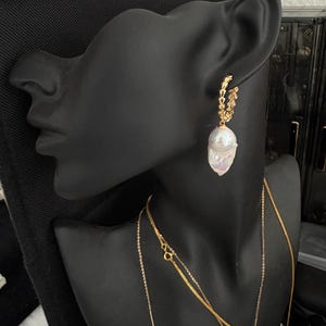 May include: A black mannequin head with a pair of gold hoop earrings with a large white pearl dangling from each earring. The mannequin is wearing two gold chain necklaces, one with a small white pearl pendant.
