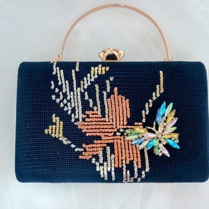 May include: A black velvet clutch with a gold top handle and a jeweled embellishment. The embellishment is a cluster of iridescent rhinestones in shades of blue, green, and pink, resembling a flower.