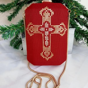 Könnte beinhalten: Eine rote Clutch mit einer goldenen Kettenkette und einem goldenen Kreuzdesign. Das Kreuz besteht aus Pailletten und Perlen.
