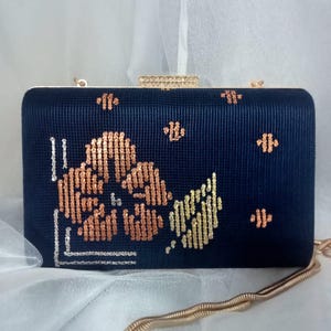 Può includere: Una pochette blu navy con una tracolla a catena dorata. La borsa è decorata con un motivo floreale in tonalità arancio e oro.