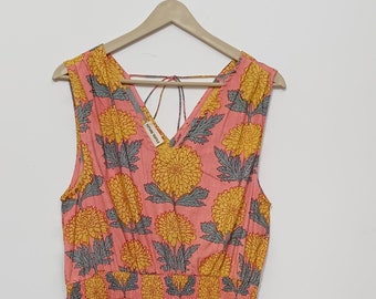 May include: A sleeveless, midi-length dress with a pink background and a repeating pattern of yellow and gray floral designs. The dress has a V-neck and a gathered waist. The dress is hanging on a wooden hanger.