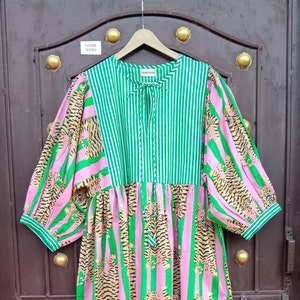 Puede incluir: Un vestido de rayas rosas y verdes con estampado de tigre. El vestido tiene una cintura fruncida y un escote redondo. El vestido está colgado de una percha de madera.