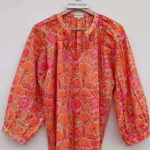 Pode incluir: Uma blusa com estampa floral em tons de laranja e rosa, pendurada em um cabide de madeira. A blusa tem mangas compridas, decote em V e borlas decorativas. O cabide tem o texto "FUTURE TEXTILE".
