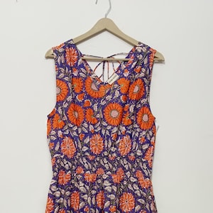 Puede incluir: Un vestido maxi sin mangas con escote en V y estampado floral. El vestido tiene una falda de capas y un lazo en la parte posterior del cuello. El estampado presenta girasoles naranjas sobre un fondo morado.