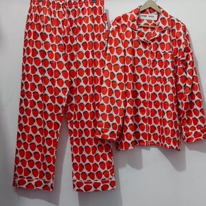 Könnte beinhalten: Ein zweiteiliges Pyjama-Set mit einem rot-weißen Erdbeermuster. Das Set besteht aus einem langärmeligen Hemd mit Brusttasche und einer Hose mit Kordelzug. Die Pyjamas hängen an einer Holzstange.