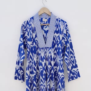 May include: A blue and white ikat print dress with a V-neck and 3/4 sleeves. The dress has a white stripe trim around the neckline. The dress is hanging on a wooden hanger.