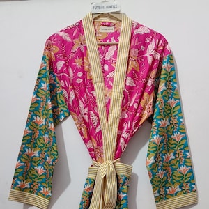 Puede incluir: Una bata con estampado floral rosa vibrante con detalles turquesa y amarillos. La bata tiene un cinturón a juego y está etiquetada como "FUTURE TEXTILE" en la percha. Las mangas tienen un estampado floral turquesa.