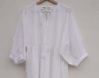 Könnte beinhalten: Ein weißes Midikleid mit kurzen Ärmeln und geraffter Taille, das an einem Holzbügel hängt. Das Kleid hat ein Bindedetail am Ausschnitt. Die Worte "FUTURE TEXTILE" sind auf einem am Bügel befestigten Etikett aufgedruckt.
