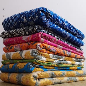 Könnte beinhalten: Ein Stapel gefalteter Stoffe in verschiedenen Farben und Mustern. Die Stoffe weisen florale, geometrische und abstrakte Designs in Blau-, Rosa-, Orange-, Gelb- und Grüntönen auf. Die Stoffe sind auf einer hellbraunen Holzoberfläche gestapelt.