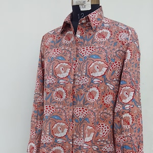 Peut inclure: Une chemise à manches longues boutonnée de couleur rose poudré, ornée d'un motif floral répétitif dans des tons de rouge, blanc et bleu. La chemise présente un col classique, une patte de boutonnage avant et des poignets boutonnés sur mannequin.